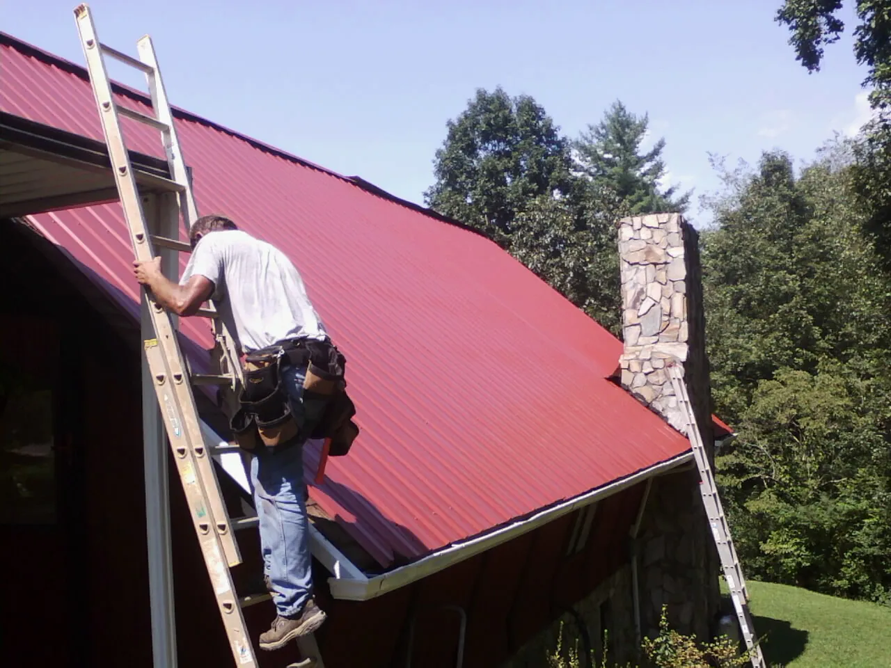 Roofer on a ladder beside a red metal roof and stone chimney — Soffit & Fascia Repair by Roofing Brothers in Coffeyville